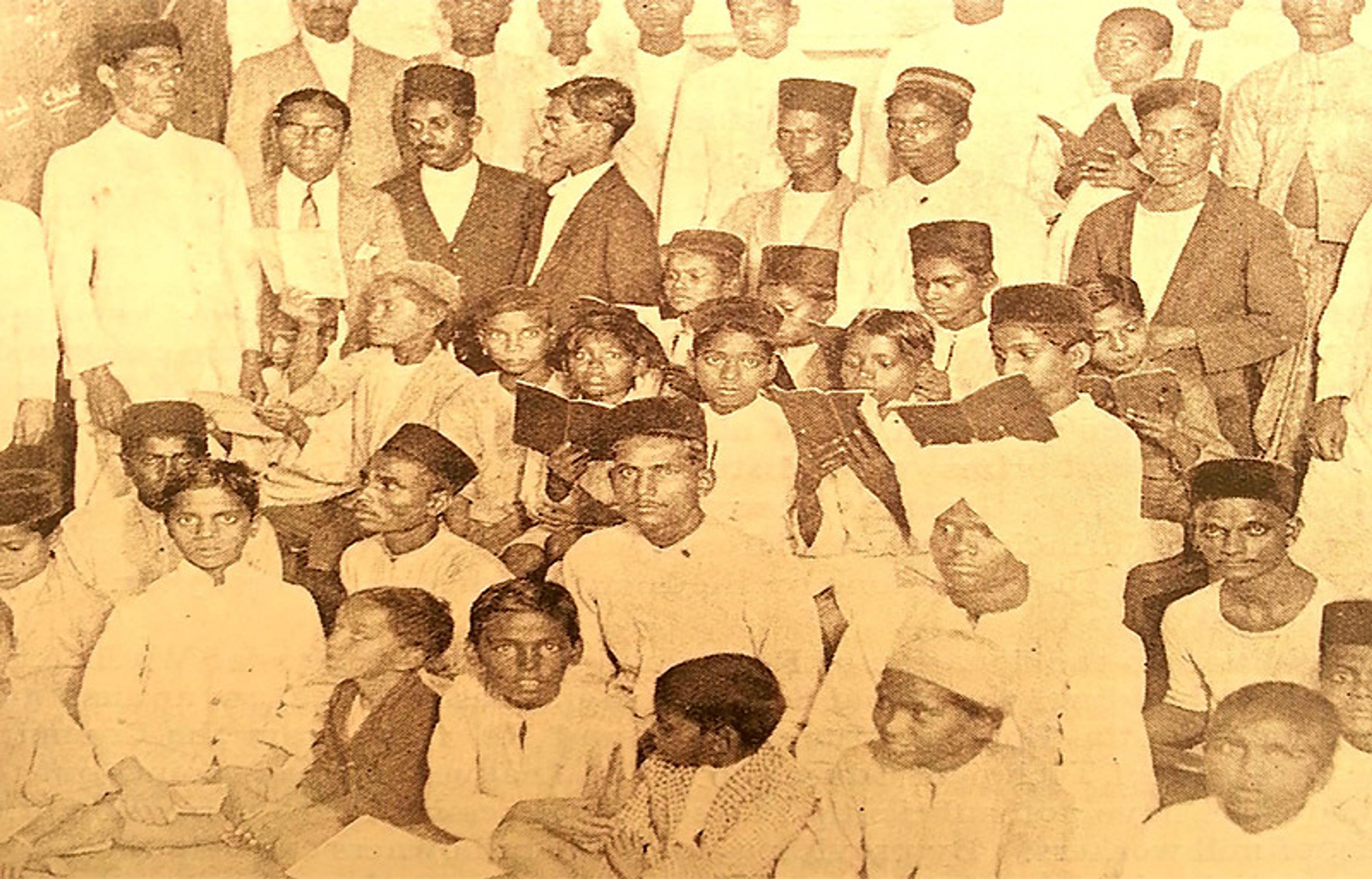 A vintage photograph showing a group of young boys and men, many wearing traditional caps and holding books, in a crowded setting.