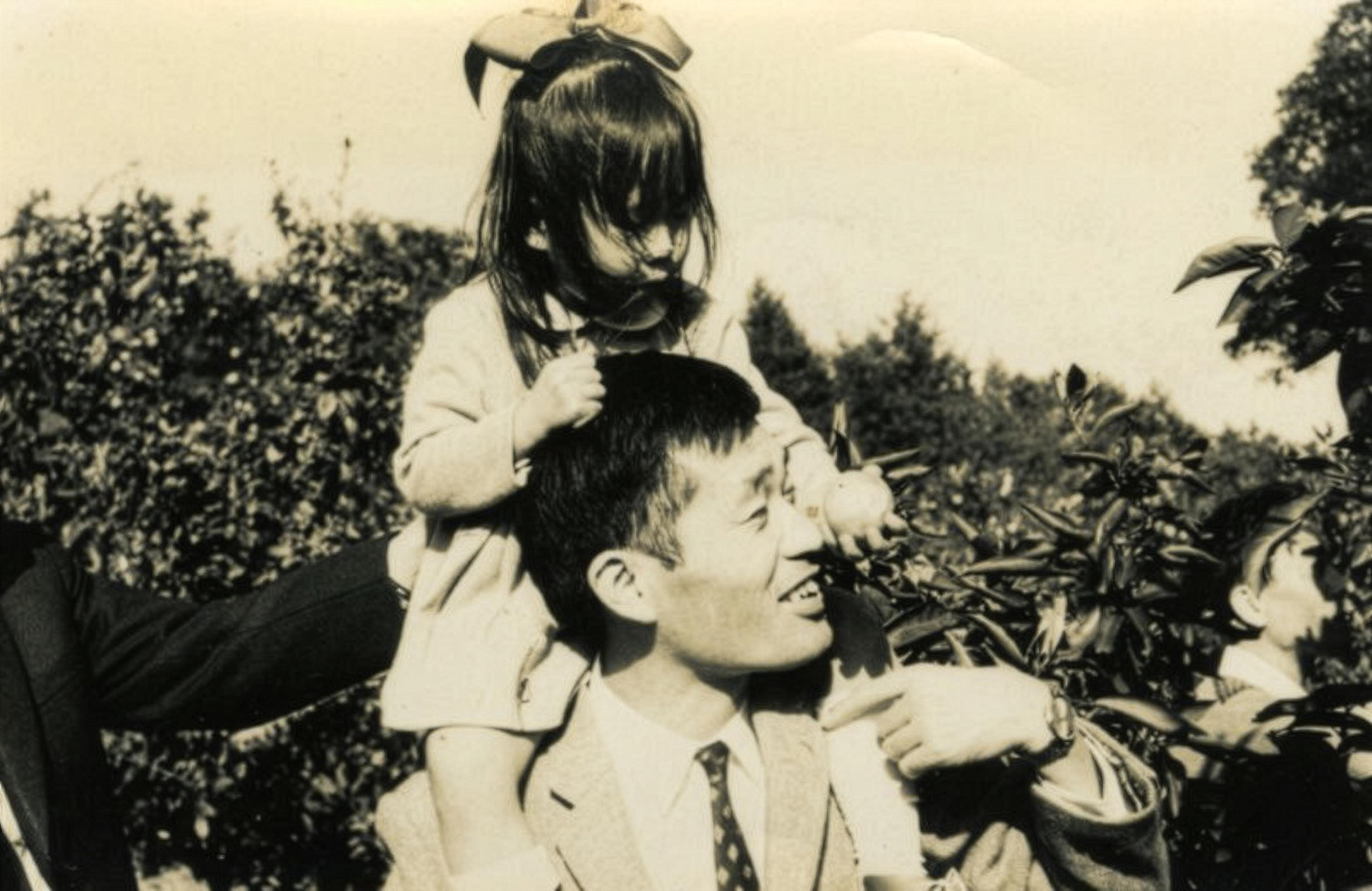Black and white photo of a man carrying a child with a bow on his shoulders in an orchard.