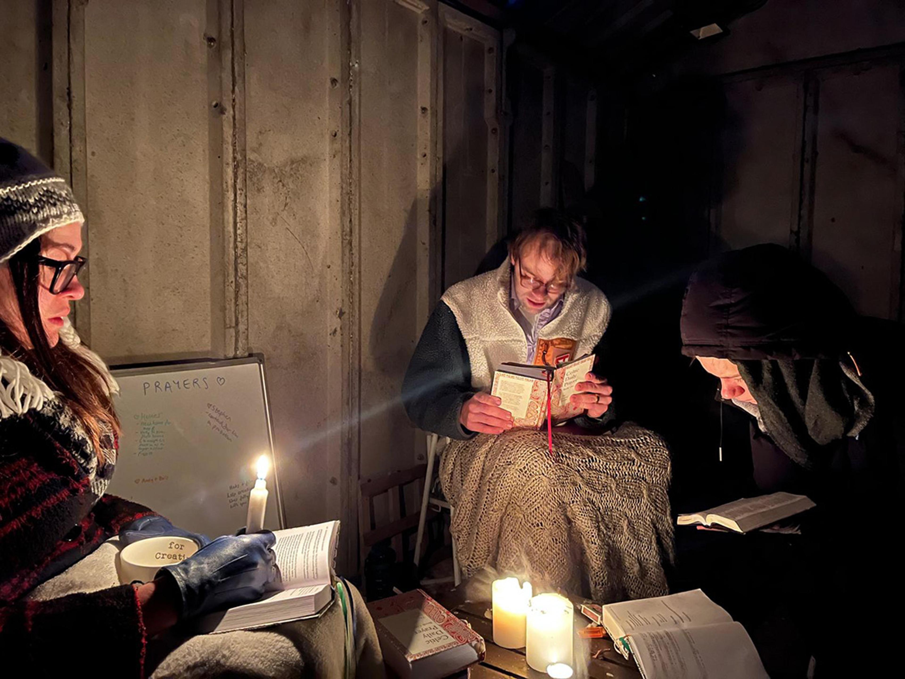 Three people wrapped in blankets and reading by candlelight in a dim room with a whiteboard labelled "Prayers".
