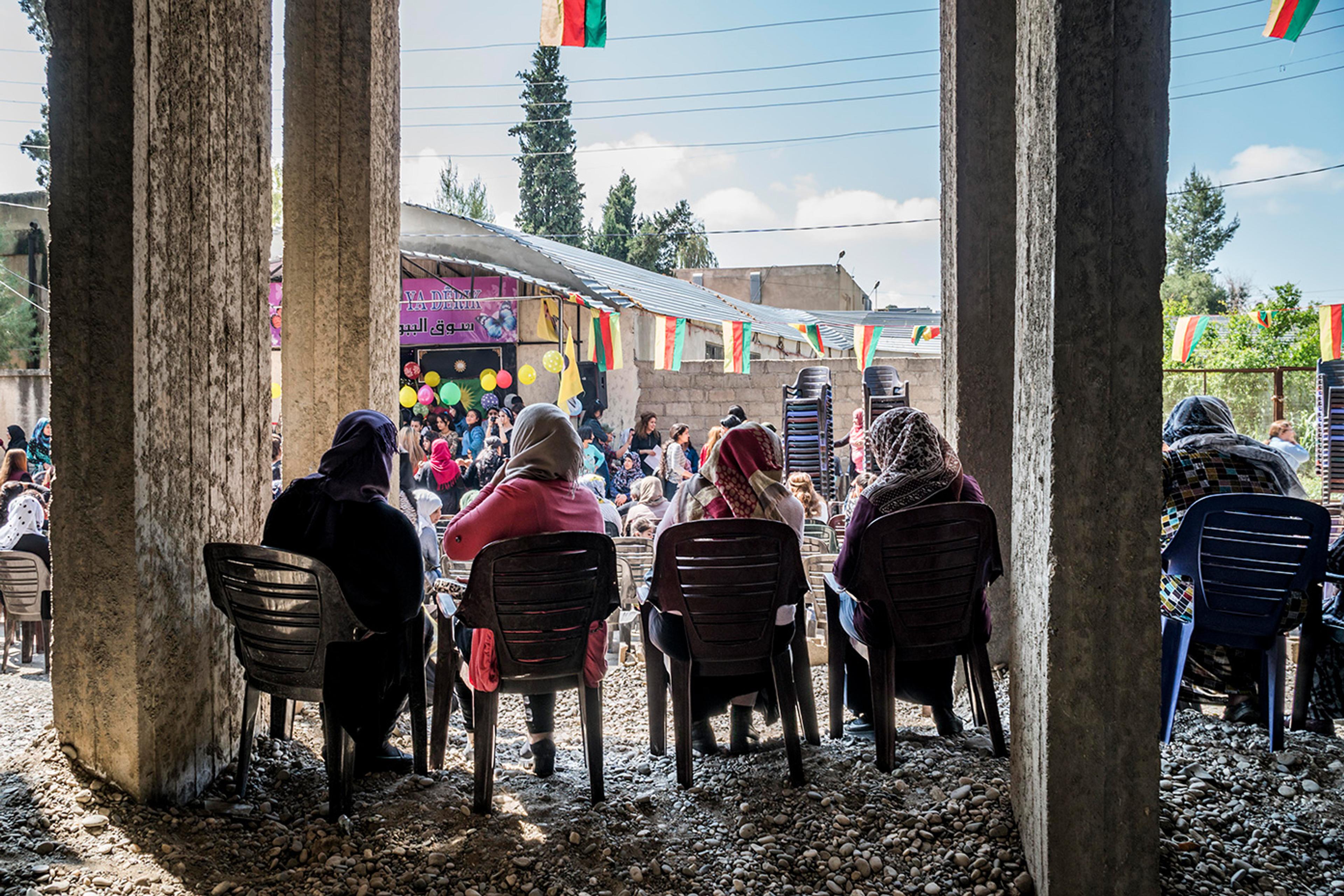 Women seated outdoors under a canopy, viewed from behind through concrete columns, with flags and balloons visible.