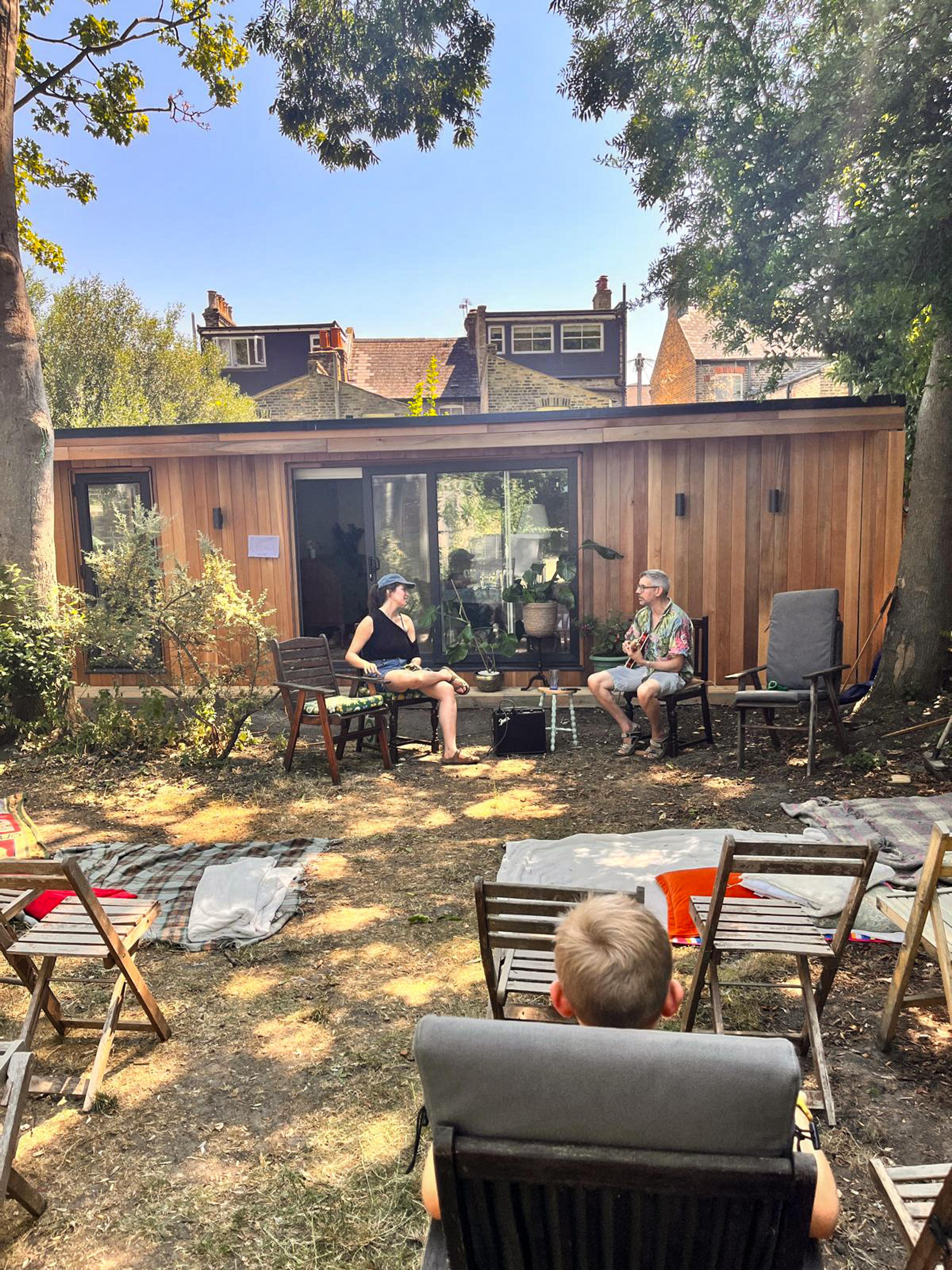 A garden gathering with people sitting outside a wooden cabin on a sunny day, surrounded by trees and chairs.