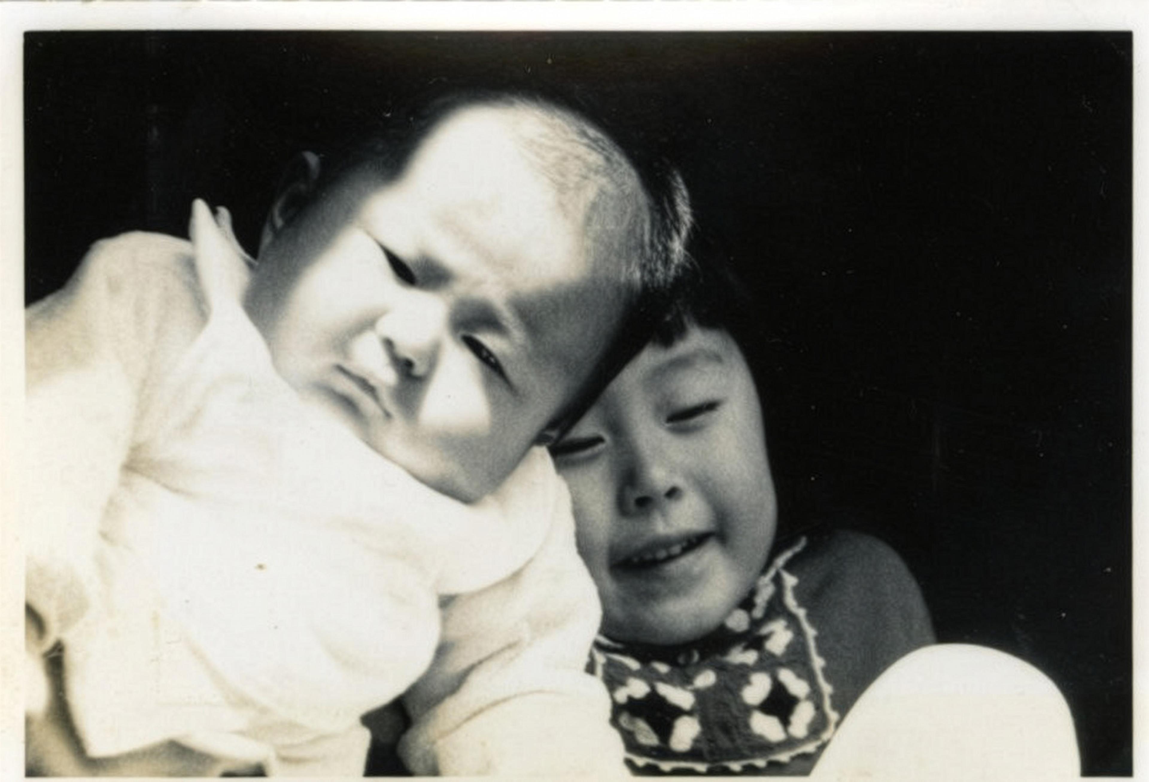 Black and white photo of a baby and a young child with a smiling face, in sunlight against a dark background.